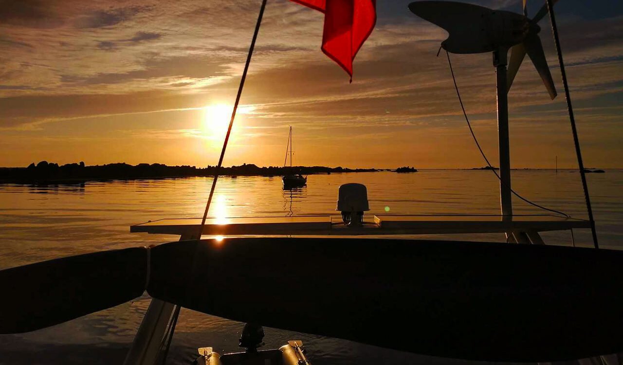 coucher de soleil au contraste de couleurs orangées et sombres depuis l'arrière d'un voilier. on aperçoit l'éolienne et les pataras.